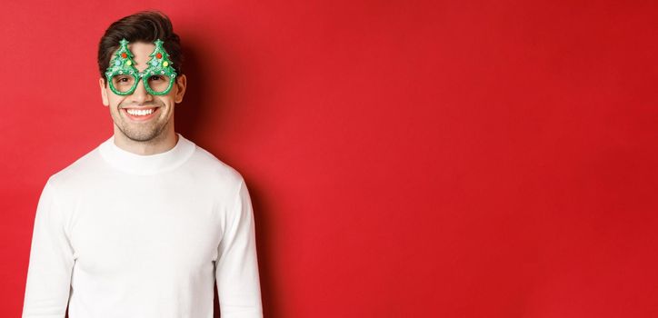 Concept of christmas, winter holidays and celebration. Close-up of handsome man in white sweater and party glasses, enjoying new year, standing over red background.