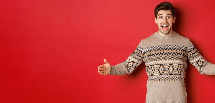 Portrait of attractive happy man in christmas sweater showing big present, spread hands sideways as if holding large box, standing over red background.