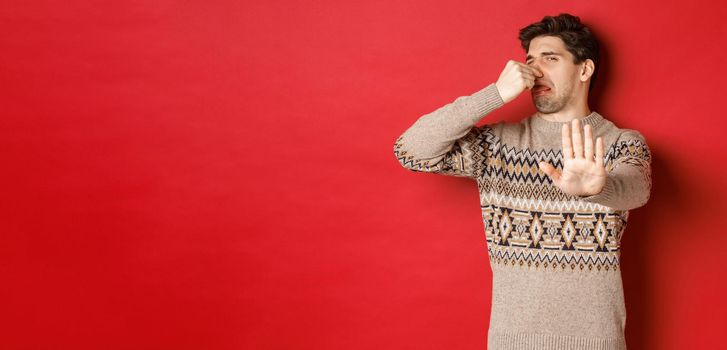 Image of disgusted handsome man in winter sweater, rejecting something with bad smell, shut nose and raising hand to decline, standing over red background.