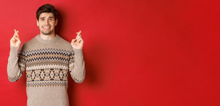 Image of hopeful and worried man in christmas sweater waiting for something, cross fingers for good luck and making wish, nervous about new year gift, red background.