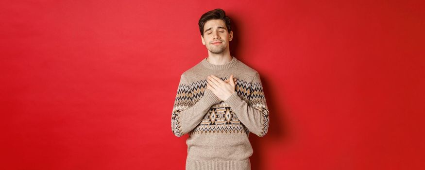 Image of dreamy and happy, attractive bearded man in christmas sweater, holding hands on heart, smiling with eyes closed, remember something touching, celebrating new year, red background.