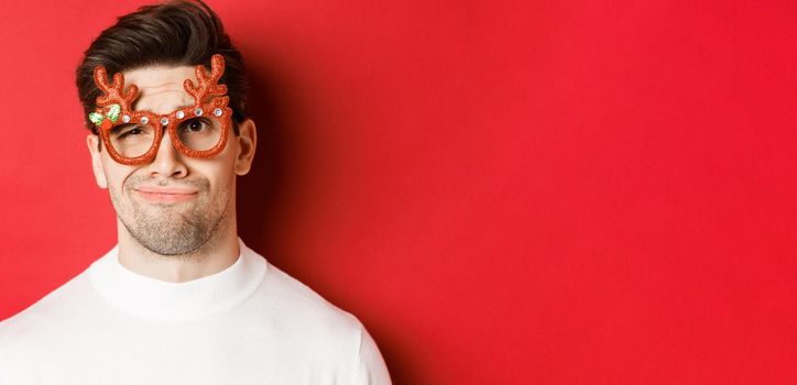 Concept of winter holidays, christmas and celebration. Close-up of skeptical handsome man in party glasses, looking doubtful and unamused, standing against red background.