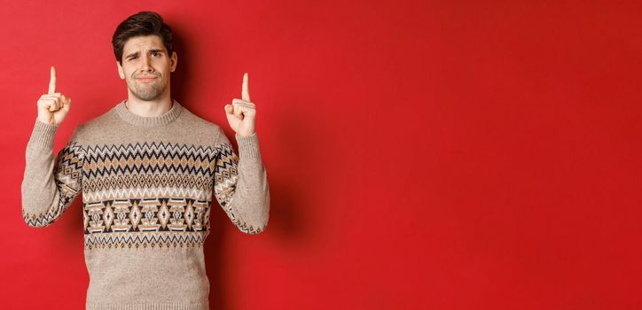 Image of skeptical and unamused handsome guy, showing bad christmas offer, pointing fingers up and grimacing displeased, express dislike, standing over red background.