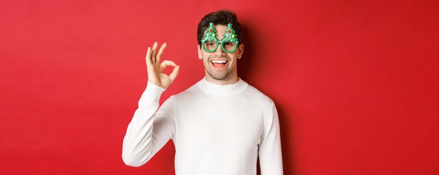 Happy caucasian guy wishing merry christmas, celebrating winter holidays, showing okay sign and smiling pleased, standing over red background.