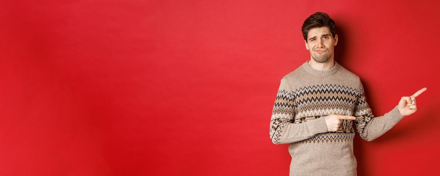 Concept of christmas celebration, winter holidays and lifestyle. Image of skeptical and displeased handsome man in xmas sweater, pointing fingers right and do not recommend product.