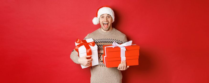 Concept of winter holidays, new year and celebration. Image of amazed and happy man in sweater and santa hat, holding christmas gifts and shouting for joy, red background.