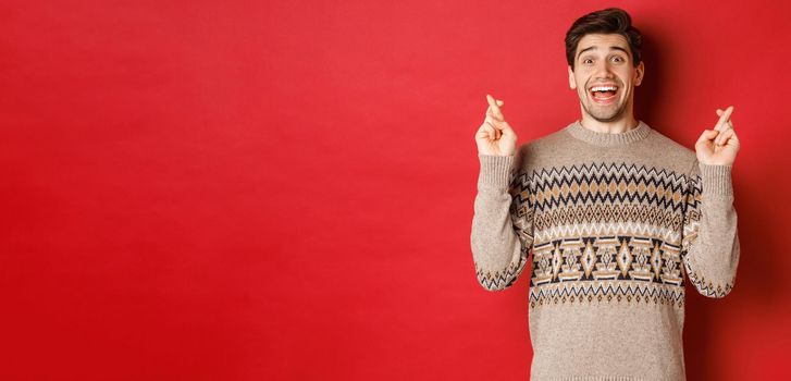 Image of excited and hopeful young man making wish, wearing winter sweater, crossing fingers for good luck and smiling, anticipating christmas eve, standing over red background.