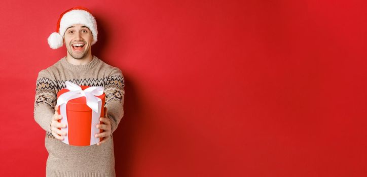 Image of happy man saying merry christmas and giving you a gift in box, smiling cheerful, celebrating winter holidays in santa hat and xmas sweater, standing over red background.