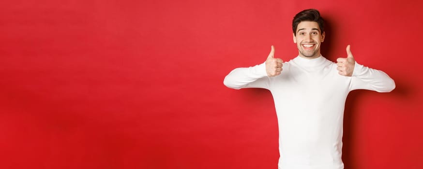 Cheerful good-looking male model in white sweater, showing thumbs-up in approval, like something good, standing over red background and smiling pleased.