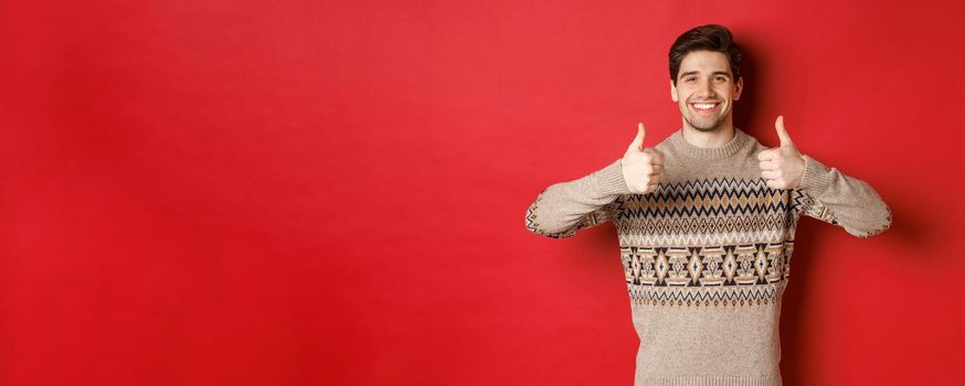 Portrait of happy and pleased handsome guy in christmas sweater, showing thumbs-up and nod in approval, smiling satisfied, standing over red background.