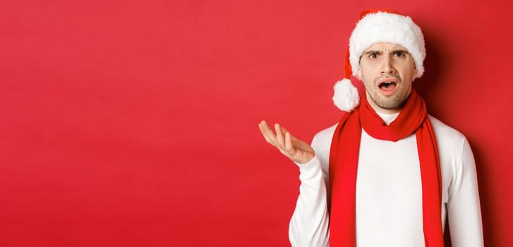 Concept of christmas, winter holidays and celebration. Portrait of confused man in santa hat and scarf, frowning and looking perplexed, standing over red background.