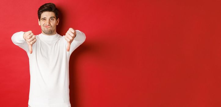 Portrait of disappointed skeptical guy, wearing white sweater, showing thumbs-down to disagree, express dislike, standing over red background.