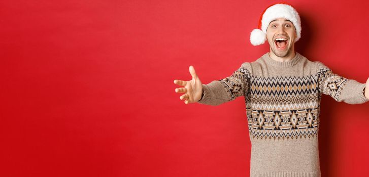 Portrait of happy, attractive man in christmas sweater and santa hat, reaching hands for gift, want to take something and smiling, standing over red background.