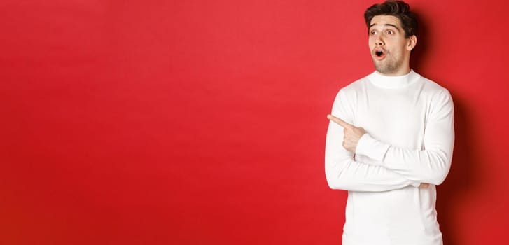 Concept of winter holidays. Portrait of impressed handsome man in white sweater, pointing and looking left with dropped jaw and amazed expression, standing against red background.
