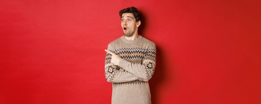 Portrait of attractive caucasian man celebrating new year, winter holidays, pointing finger and looking left surprised, showing christmas advertisement, standing over red background.