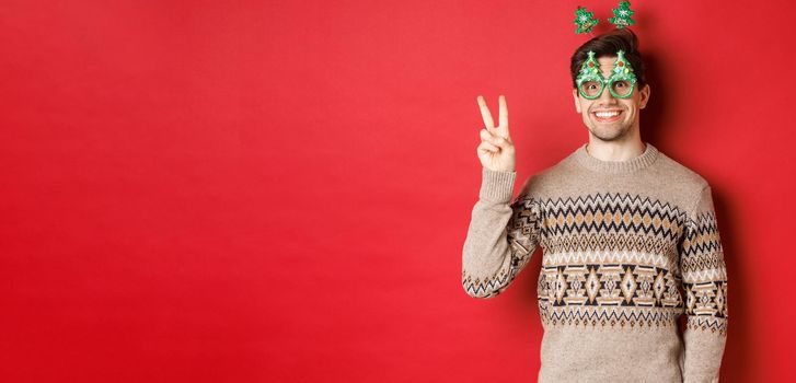 Portrait of funny and cute man in party glasses and christmas sweater, smiling and showing peace sign while posing over red background for new year advertisement.