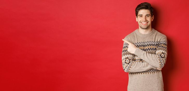 Image of handsome man in christmas sweater, celebrating new year holidays, smiling happy and pointing finger at upper left corner copy space, standing over red background.