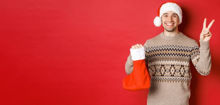 Concept of winter holidays, new year and celebration. Image of happy smiling man in santa hat and sweater, showing peace sign and a christmas stocking bag with gifts, red background.