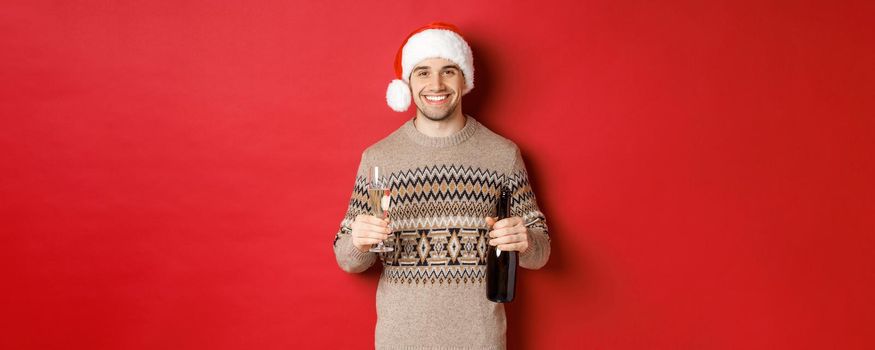 Concept of winter holidays, new year and celebration. Portrait of handsome man in santa hat and sweater, holding champagne and making toast on christmas party, standing over red background.