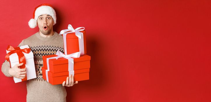 Concept of winter holidays, new year and celebration. Image of surprised handsome guy in sweater and santa hat, receiving gifts, holding boxes with presents and looking amazed.