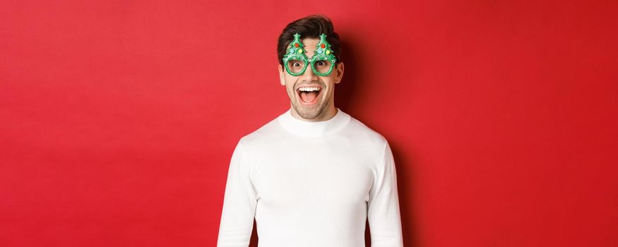 Concept of christmas, winter holidays and celebration. Close-up of handsome young man enjoying new year, wearing party glasses and looking amazed at camera, standing over red background.