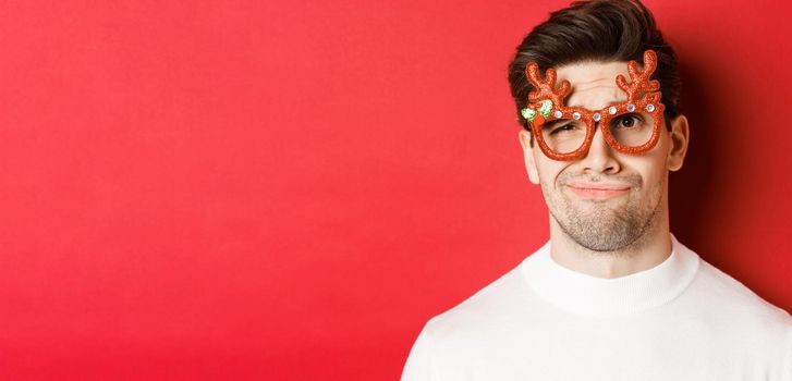 Concept of winter holidays, christmas and celebration. Close-up of skeptical handsome man in party glasses, looking doubtful and unamused, standing against red background.