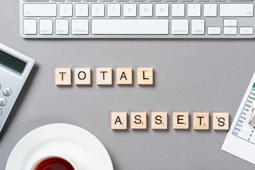 Total assets concept with letters on wooden cubes. Still life of office workplace with supplies. Flat lay grey surface with computer keyboard and cup of tea. Capital investment and brokerage company.