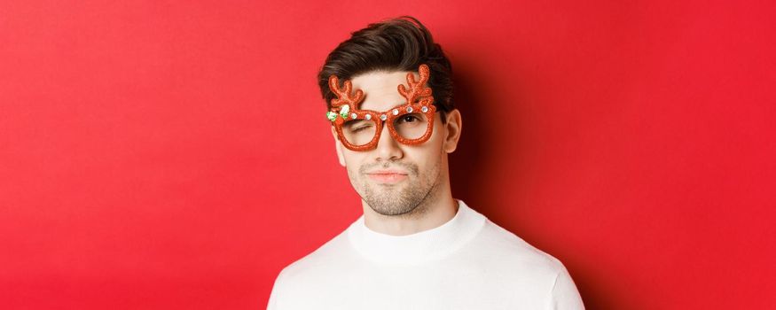 Concept of winter holidays, christmas and celebration. Close-up of cheeky attractive man in party glasses, winking and looking sassy, standing over red background.