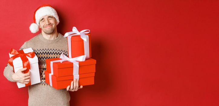 Concept of winter holidays, new year and celebration. Portrait of lovely smiling man receiving pile of presents, holding gifts and being touched with surprise, standing over red background grateful.