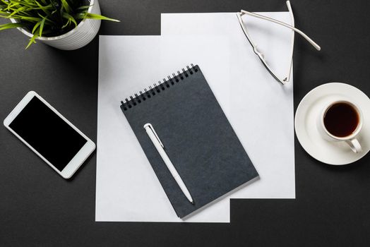 Top view of modern workplace with spiral notepad. Flat lay black surface with smartphone and cup of coffee. Top view coworking workspace and freelance. Digital technology and mobile lifestyle.