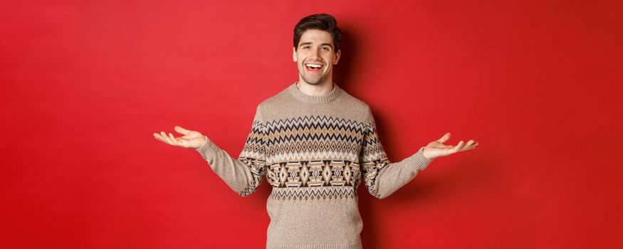 Portrait of happy good-looking man celebrating new year holidays, wearing christmas sweater, spread hands sideways and smiling, holding something on copy space, standing over red background.