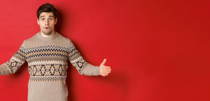 Image of amazed handsome man showing size of christmas present, spread hands sideways and shaping something large, standing over red background.