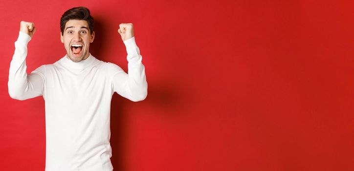 Portrait of excited lucky man in white sweater, raising hands up and triumphing, celebrating new year, standing happy against red background.