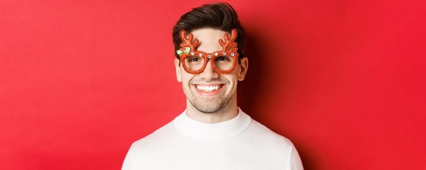 Concept of winter holidays, christmas and celebration. Close-up of attractive brunette guy in party glasses, smiling and looking happy, standing against red background.