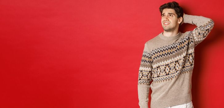 Image of puzzled and indecisive man in christmas sweater, looking at upper left corner, thinking about new year holidays gifts, standing over red background.