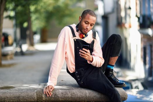 Young black man with headphones sitting in urban street looking at his smart phone. Lifestyle concept.