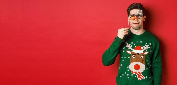 Funny man in christmas sweater and party mask, celebrating winter holidays, showing funny faces, standing over red background.