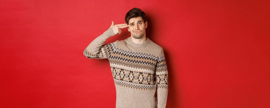 Portrait of distressed and annoyed handsome man, wearing christmas sweater, making finger gun sign near head and shooting himself, standing over red background.