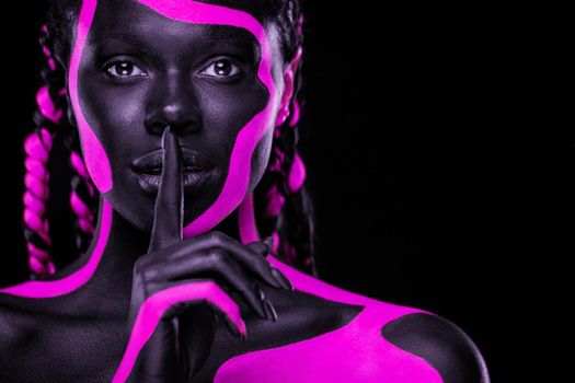 Face art. Girl showing gesture quietly, asks remain silent with finger. Woman with black and pink body paint. Young african girl with colorful bodypaint. An amazing afro american model with makeup