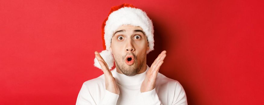 Close-up of amazed handsome man in santa hat and white sweater, raising hands and looking impressed, standing over red background.