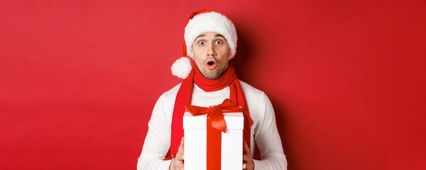 Concept of winter holidays, christmas and lifestyle. Close-up of surprised handsome guy in santa hat and scarf, looking amazed and holding new year gift, standing over red background.