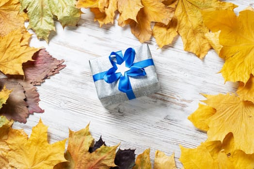 Flat lay autumn composition with gift box and yellow autumn leaves. Holiday present decorated blue ribbon bow on vintage wooden desk. Happy thanksgiving congratulation. Autumn sale advertising.