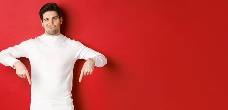 Portrait of attractive, skeptical young man in white sweater, grimacing and pointing fingers down, disagree and dislike something, standing over red background.