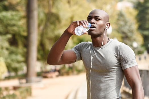Young black man drinking water before running in urban background. Young male exercising with naked torso listening to music with headphones.