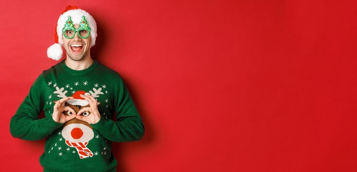 Portrait of carefree handsome man in santa hat and party glasses, making fun of his christmas sweater, looking happy over red background.