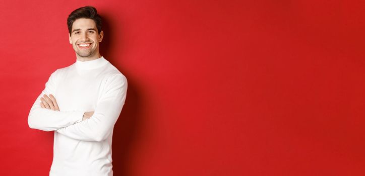 Concept of winter holidays, christmas and lifestyle. Portrait of confident attractive guy in white sweater, smiling with hands crossed on chest, standing over red background.