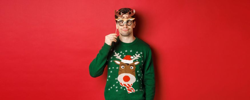 Funny man in christmas sweater and party mask, celebrating winter holidays, showing funny faces, standing against red background.