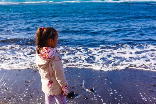 Little girl wearing jacket is playing at the beach ocean on cloudy sky in winter time