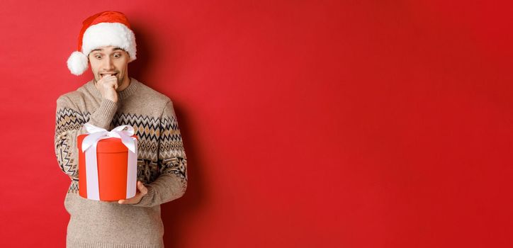 Image of worried guy in santa hat and sweater, looking indecisive at christmas gift, standing against red background.