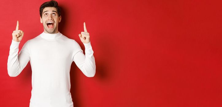 Concept of winter holidays. Image of amazed handsome guy reacting to christmas announcement, pointing and looking up at copy space, standing over red background.